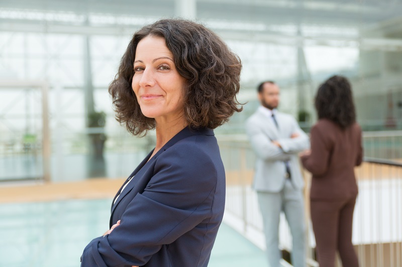 Friendly successful business woman posing with arms crossed, looking at camera, smiling. Her male and female colleagues standing and talking in background. Female business leader concept